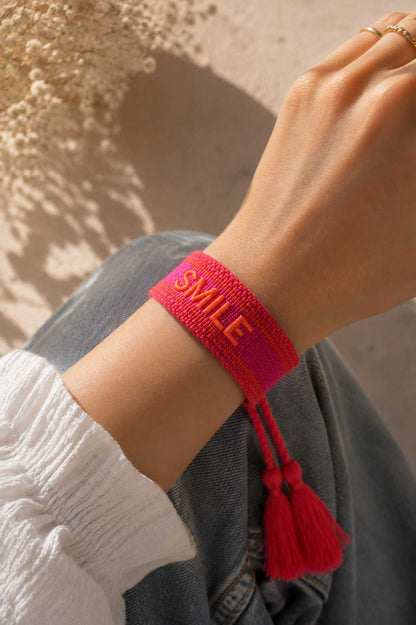 Pinkes SMILE Armband mit roten Quasten am Handgelenk getragen, weißes Oberteil und Jeans, Sonnenlicht – Saltyblush
