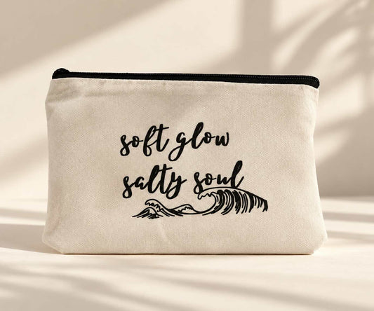 Beige Kosmetiktasche mit schwarzem Reißverschluss, Schriftzug „soft glow salty soul“ und gezeichnetem Wellenmotiv auf hellem Untergrund im Sonnenlicht.
