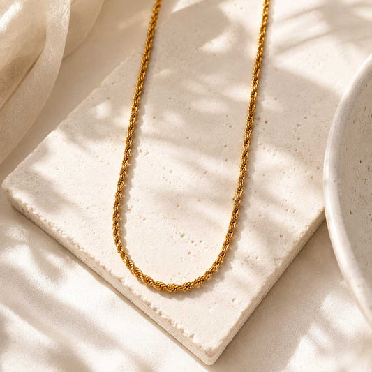 Goldene Rope-Halskette auf weißem Stein mit cremefarbenem Seidenstoff im Sonnenlicht, vergoldete Kordelkette aus Edelstahl mit gedrehtem Muster
