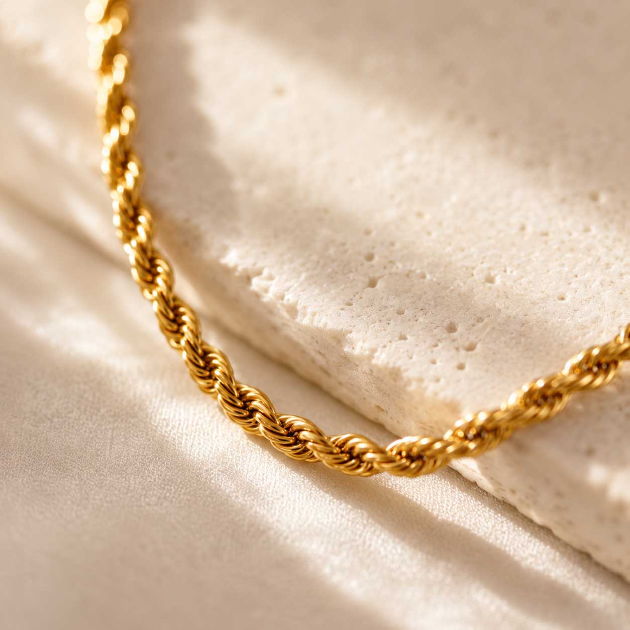 Nahaufnahme goldener Rope-Halskette auf weißem Stein, Detailansicht des gedrehten Kordelmusters der vergoldeten Edelstahlkette im Sonnenlicht
