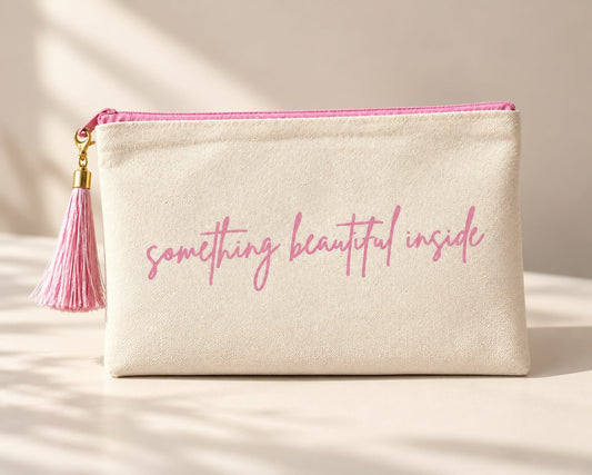 Beige Kosmetiktasche mit rosa Schriftzug „something beautiful inside“, rosa Reißverschluss und rosa Quaste mit goldfarbenen Details auf hellem Untergrund.
