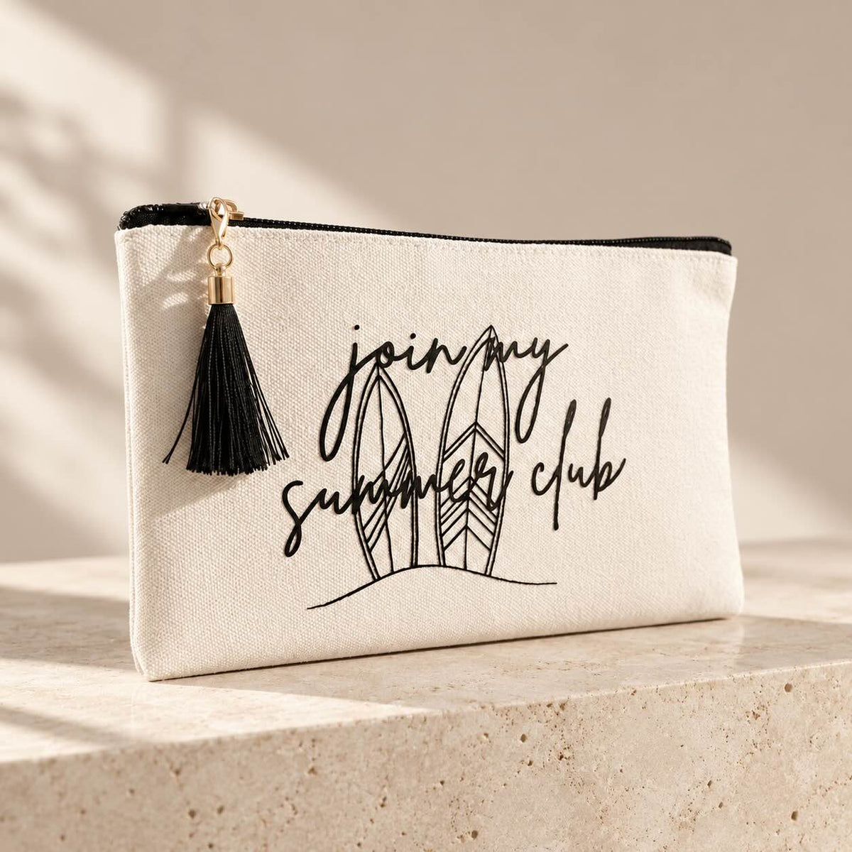 Beige summer club Kosmetiktasche mit schwarzem Reißverschluss, schwarzer Quaste und Surfbrettmotiv mit Schriftzug „join my summer club“ auf einer hellen Steinplatte.
