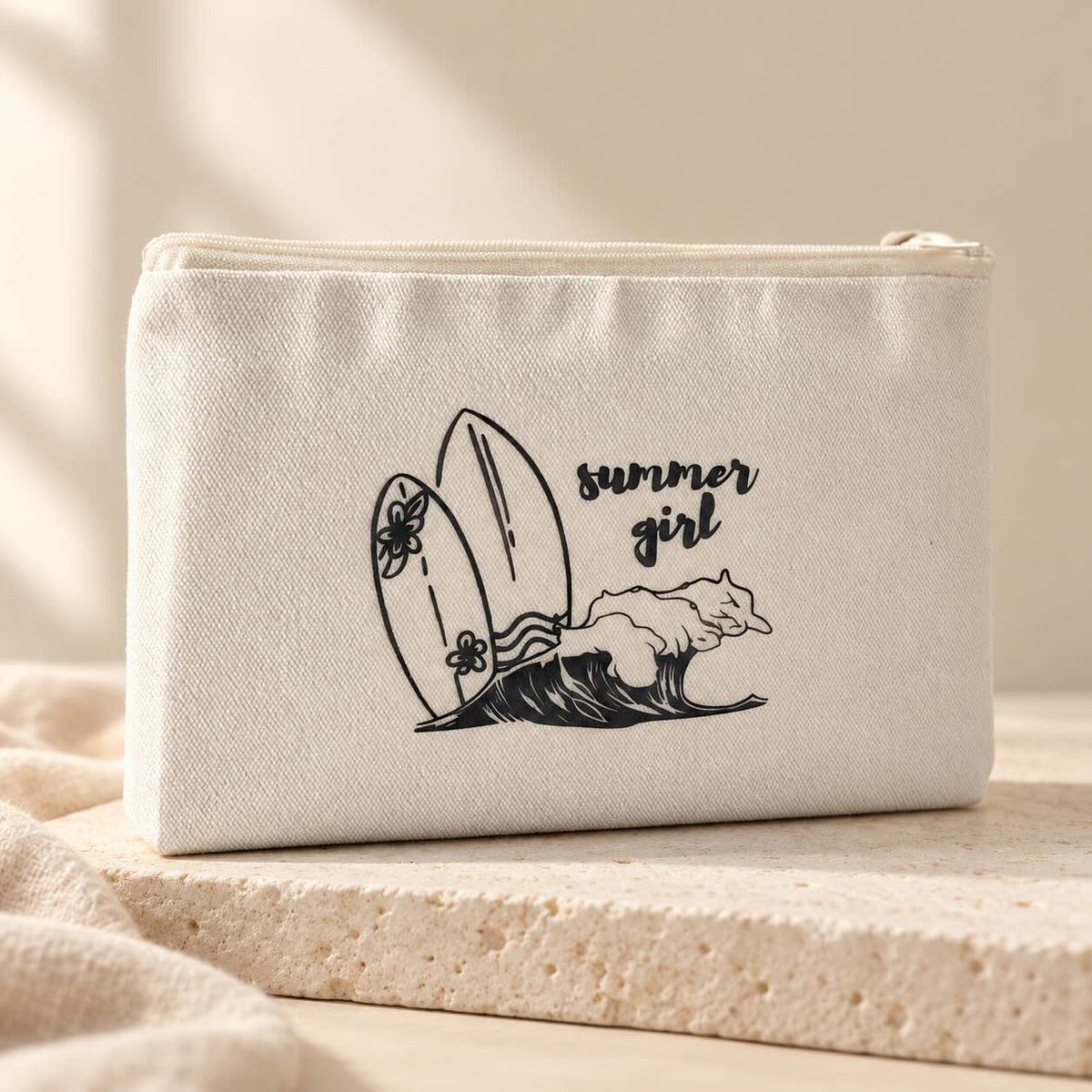 Beige summer girl Kosmetiktasche mit Reißverschluss und schwarzem Aufdruck aus zwei Surfbrettern, einer Welle und dem Schriftzug „summer girl“ auf einer hellen Steinplatte.
