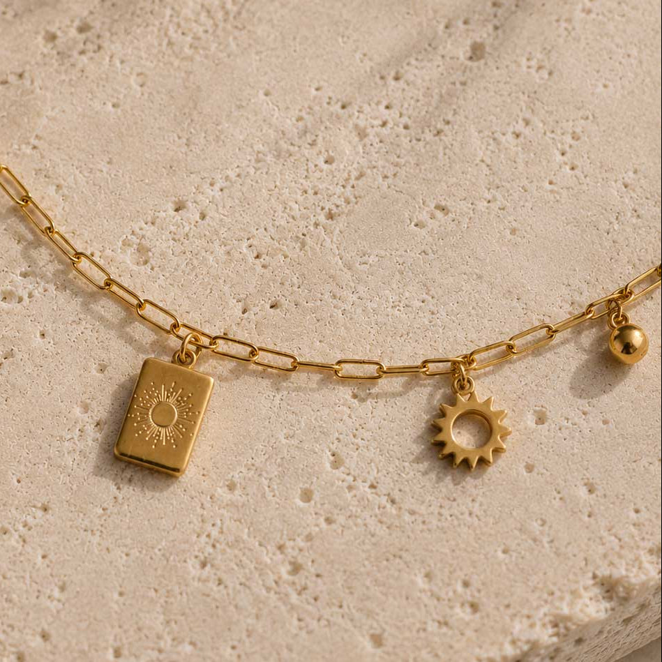 Nahaufnahme der Sundrop goldenen Fußkette aus Edelstahl mit Sonnen-Medaillon, Sonnenrad-Charm, Perle und Goldkugel auf Travertin – Saltyblush
