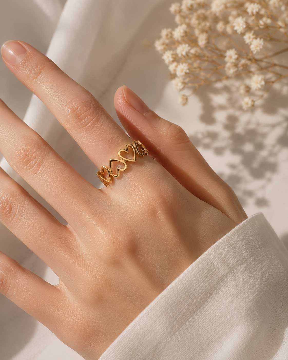 Goldener Herzring am Finger getragen, größenverstellbarer Ring mit Herz-Muster und Schleierkraut im Hintergrund
