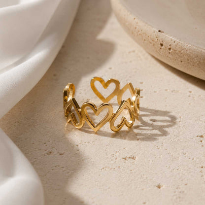Goldener Ring mit Herz-Motiven auf Sandstein-Untergrund, vergoldeter offener Herzring mit weißem Stoff
