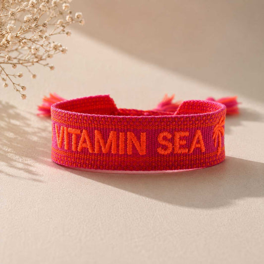 Rotes Statement Armband mit oranger Vitamin Sea Stickerei und Palmen-Motiv, rote Quasten, Trockenblumen im Hintergrund – Saltyblush
