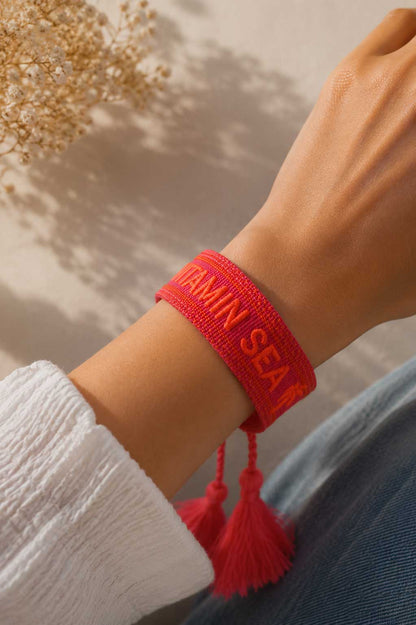 Rotes Vitamin Sea Armband mit roten Quasten am Handgelenk getragen, weißes Oberteil, Trockenblumen im Hintergrund – Saltyblush
